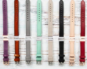 May include: A selection of watch straps in a range of colours: purple, brown, grey, green, beige, black, nude, and red. Each strap is displayed with its colour name above. The straps are leather and have a buckle fastening.