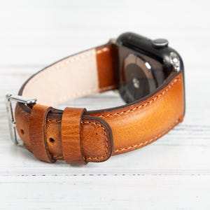 May include: A close-up of a brown leather watch band with a silver buckle. The band is a warm, medium brown color with visible stitching and a lighter interior. The watch face is black.