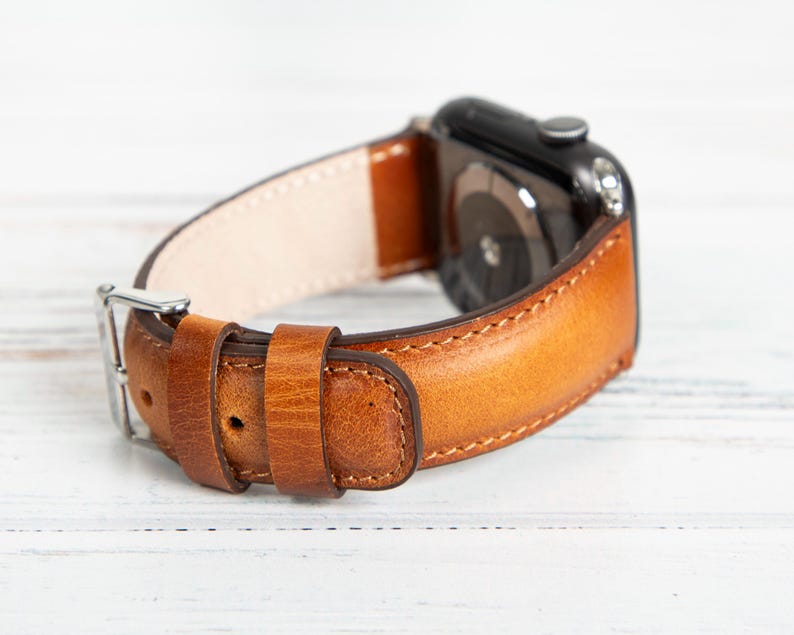 May include: A close-up of a brown leather watch band with a silver buckle. The band is a warm, medium brown color with visible stitching and a lighter interior. The watch face is black.