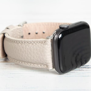 May include: A light beige leather Apple Watch band with a black watch face. The band has a textured surface and a silver-colored clasp. The watch face is black and has a silver-colored button.