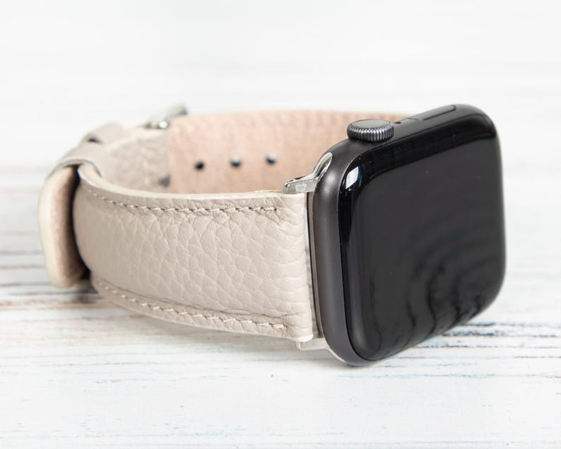 May include: A light beige leather Apple Watch band with a black watch face. The band has a textured surface and a silver-colored clasp. The watch face is black and has a silver-colored button.