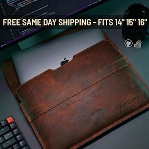 May include: A brown leather laptop sleeve with a laptop inside. The sleeve has the text "<DEVELOPER /> github.com/USERNAME" and fits 14", 15", and 16" laptops. A black keyboard and mug are in the background.