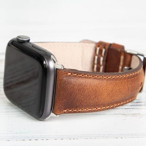 May include: A close-up of a smart watch with a brown leather band. The watch face is black, and the band is stitched with white thread. The band has a silver buckle.