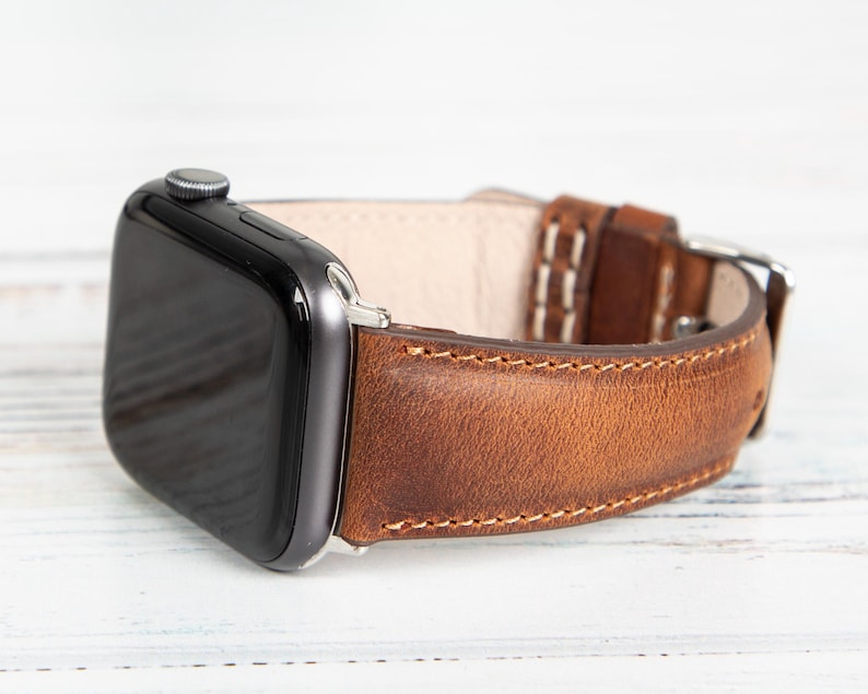 May include: A close-up of a smart watch with a brown leather band. The watch face is black, and the band is stitched with white thread. The band has a silver buckle.