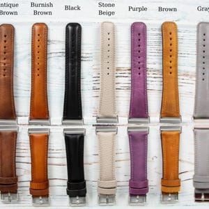 May include: A collection of leather watch bands in various colors, including antique brown, burnish brown, black, stone beige, purple, brown, and gray. Each band has a silver-colored buckle and is displayed on a light-colored wooden surface.