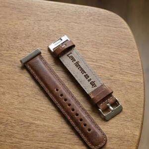 May include: Two brown leather watch straps with silver-tone hardware are displayed on a wooden surface. One strap is unattached, while the other is buckled and features the engraved text "Love forever an a day."