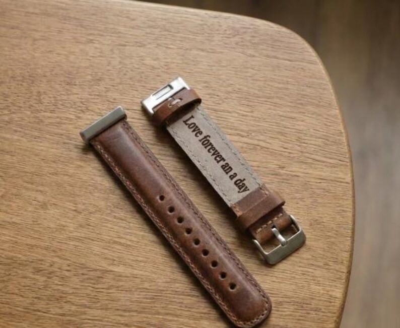 May include: Two brown leather watch straps with silver-tone hardware are displayed on a wooden surface. One strap is unattached, while the other is buckled and features the engraved text "Love forever an a day."