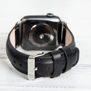 May include: Black leather watch band with a silver buckle. The band is attached to a black smartwatch. The watch band is made of smooth leather and has a classic design.
