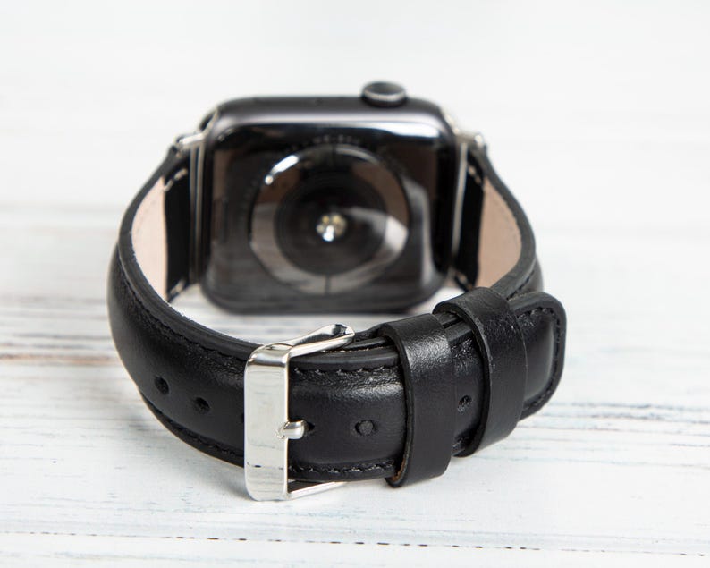 May include: Black leather watch band with a silver buckle. The band is attached to a black smartwatch. The watch band is made of smooth leather and has a classic design.