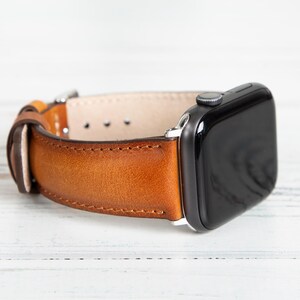 May include: A brown leather watch band with a stainless steel buckle is attached to a black smartwatch. The watch band is made of a rich, textured leather and is secured with a silver-colored buckle.