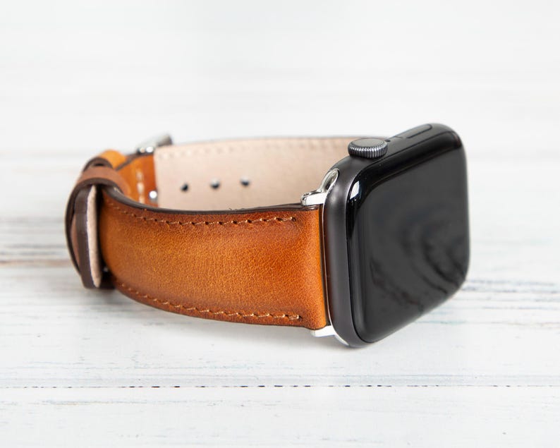 May include: A brown leather watch band with a stainless steel buckle is attached to a black smartwatch. The watch band is made of a rich, textured leather and is secured with a silver-colored buckle.