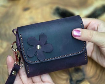 May include: A dark purple leather wallet with a flower applique and a gold-tone snap. The wallet has a wrist strap and gold-tone hardware. The stitching is a contrasting light brown color.