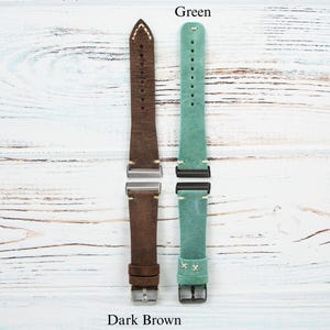 May include: Two leather watch straps, one dark brown and one green, are displayed against a white wood grain background. The dark brown strap has cream stitching and a silver buckle. The green strap has black hardware and white stitching.
