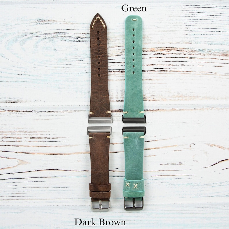 May include: Two leather watch straps, one dark brown and one green, are displayed against a white wood grain background. The dark brown strap has cream stitching and a silver buckle. The green strap has black hardware and white stitching.