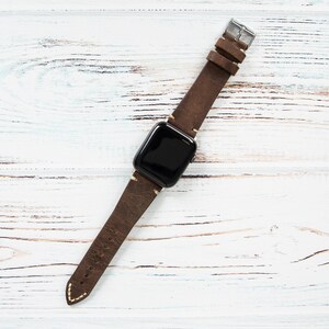 May include: A brown leather watch band with a silver buckle and a black watch face. The band is made of distressed leather and has white stitching. The watch is displayed on a white wooden surface.
