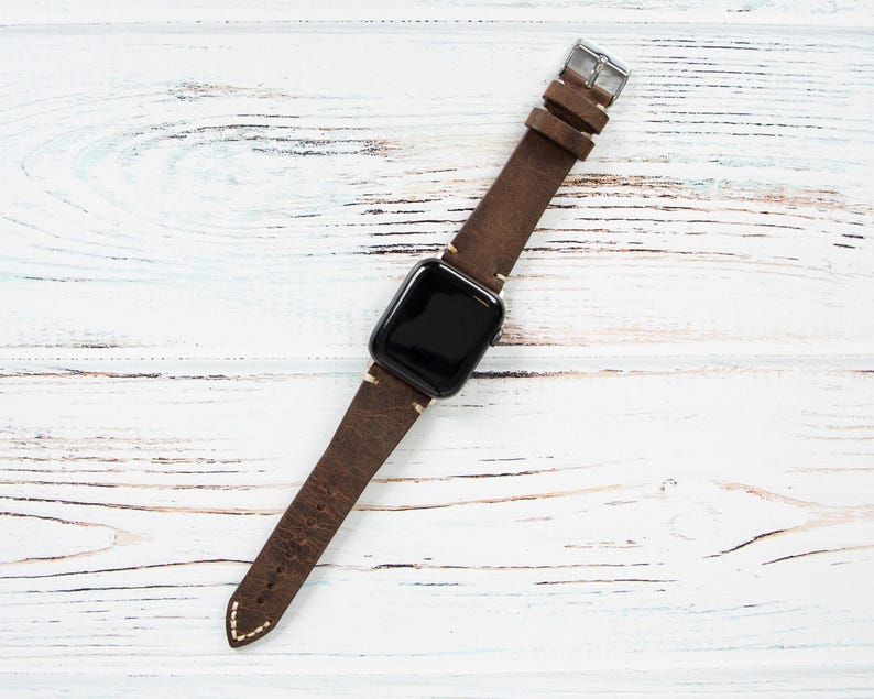 May include: A brown leather watch band with a silver buckle and a black watch face. The band is made of distressed leather and has white stitching. The watch is displayed on a white wooden surface.