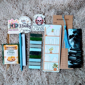 May include: A collection of book-themed accessories, including stickers, bookmarks, pens, and tea. The stickers feature text like "Books Chickens" and "Trigger Warning." A "Reading List" bookmark and a blue tassel are also present.