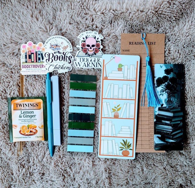 May include: A collection of book-themed accessories, including stickers, bookmarks, pens, and tea. The stickers feature text like "Books Chickens" and "Trigger Warning." A "Reading List" bookmark and a blue tassel are also present.