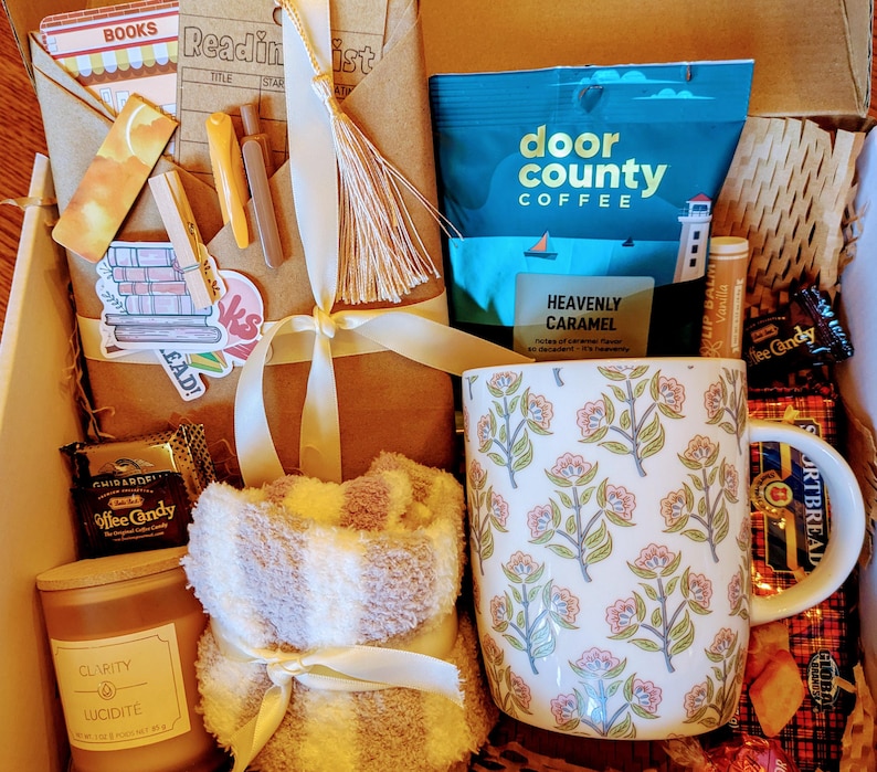 May include: A gift box filled with items for a cozy night in, including a mug with a floral pattern, a soft blanket, a candle, coffee, and candy. The box is decorated with a brown paper bag and a white ribbon.