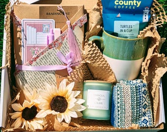 May include: A gift box containing a Door County Coffee bag, a two-toned green and white ceramic mug, a candle labeled Tranquil Calme, a pair of patterned socks, two artificial sunflowers, a reading list card, and a floral bookmark with a tassel.