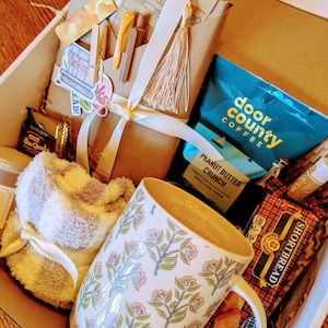 May include: A gift box filled with various items, including a mug with a floral pattern and the text "you belong among the wildflowers", a fuzzy white slipper, a bag of coffee, a bag of peanut butter crunch, and shortbread cookies.