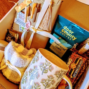 May include: A gift box filled with items for a cozy night in, including a mug with the text "you belong among the wildflowers", a fuzzy slipper sock, a book, coffee, and shortbread cookies.