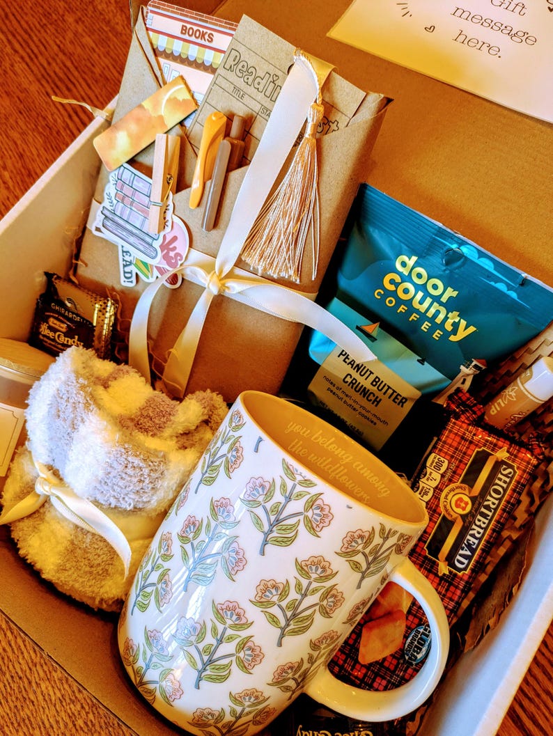 May include: A gift box filled with items for a cozy night in, including a mug with the text "you belong among the wildflowers", a fuzzy slipper sock, a book, coffee, and shortbread cookies.