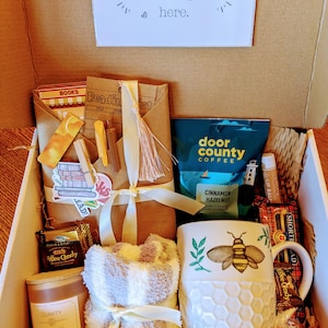 May include: A gift box filled with items for a cozy night in, including a mug with a bee design, a candle, a throw blanket, coffee, and chocolate.