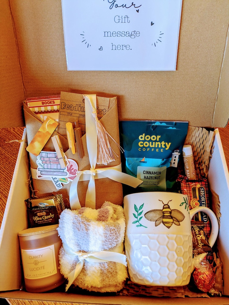 May include: A gift box filled with items for a cozy night in, including a mug with a bee design, a candle, a throw blanket, coffee, and chocolate.
