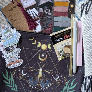 May include: A collection of reading accessories and stationery items. Includes bookmarks, stickers, a pen, a lip balm, and a tea bag. The items are arranged on a patterned surface, with a dark pouch featuring a moth and moon design.