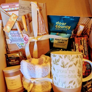 May include: A gift box filled with items for a cozy night in, including a mug that says "you're a cu-tea", a candle, a book, coffee, and a pair of socks.