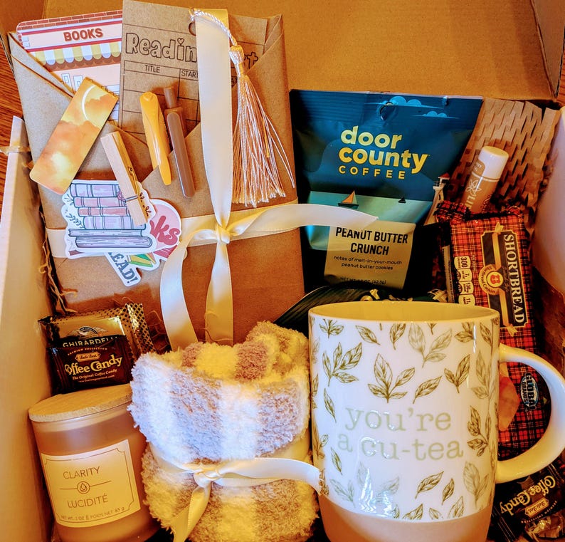May include: A gift box filled with items for a cozy night in, including a mug that says "you're a cu-tea", a candle, a book, coffee, and a pair of socks.