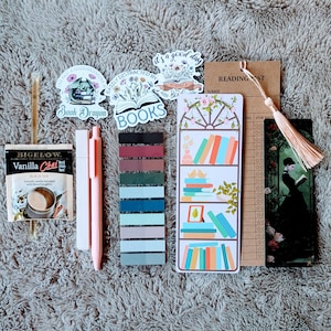 May include: A collection of book-themed accessories, including stickers, bookmarks, sticky notes, and pens. The image features a Bigelow Vanilla Chai tea bag, a reading list card, and a bookmark with a woman reading. The stickers have text that reads "Book Dragon" and "BOOKS".