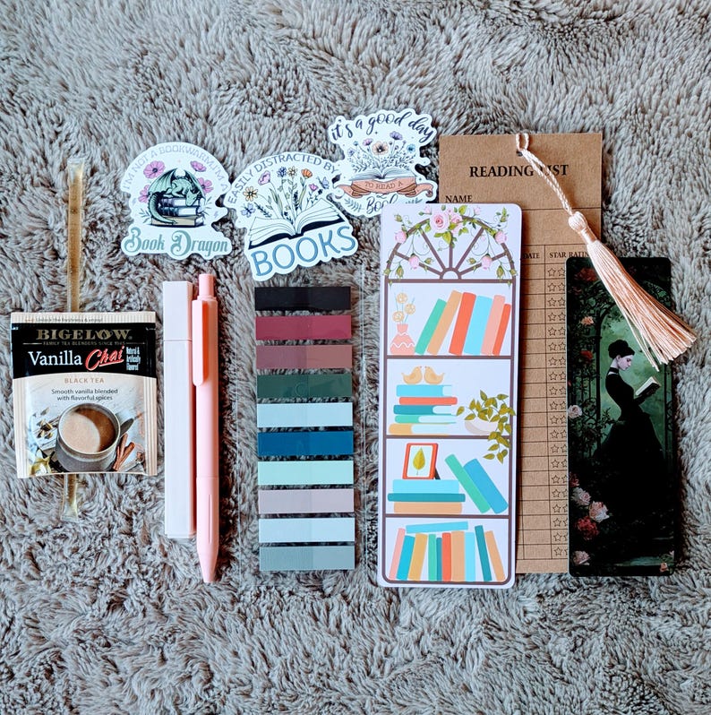 May include: A collection of book-themed accessories, including stickers, bookmarks, sticky notes, and pens. The image features a Bigelow Vanilla Chai tea bag, a reading list card, and a bookmark with a woman reading. The stickers have text that reads "Book Dragon" and "BOOKS".