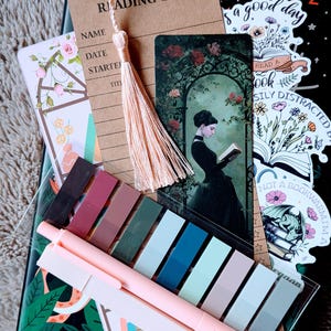 May include: A collection of stationery items for readers, including a reading list tag with a tassel, bookmarks, sticky notes, and pens. The image features a black notebook with floral designs and stickers with text like "Read a Book" and "Not a Bookworm".