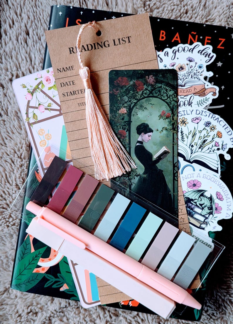 May include: A collection of stationery items for readers, including a reading list tag with a tassel, bookmarks, sticky notes, and pens. The image features a black notebook with floral designs and stickers with text like "Read a Book" and "Not a Bookworm".