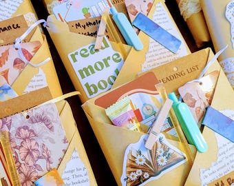May include: A collection of wrapped gifts, each in a brown paper envelope, adorned with decorative elements like lace, stickers, and small wooden clothespins. Some envelopes have the words "Read More Books" printed on them. The theme is book-related.