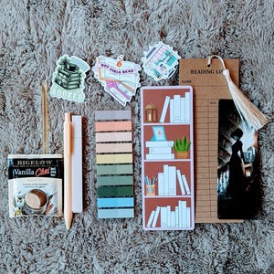 May include: An assortment of stationery items for readers, including stickers, a bookmark, sticky notes, a pen, and a tea bag. The stickers feature book-related designs and the text "BOOKED" and "HOT GIRLS READ". The bookmark has a tassel.