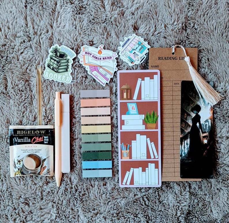 May include: An assortment of stationery items for readers, including stickers, a bookmark, sticky notes, a pen, and a tea bag. The stickers feature book-related designs and the text "BOOKED" and "HOT GIRLS READ". The bookmark has a tassel.
