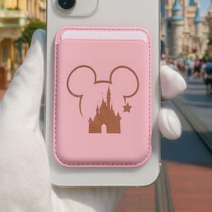 May include: A pink phone wallet with a brown Mickey Mouse silhouette and castle design is attached to a white smartphone. The background features a sunny day with a castle and buildings.