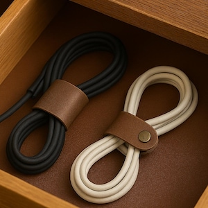 May include: Two coiled charging cables, one black and one white, are neatly organized in a wooden drawer. Each cable is secured with a brown leather strap. The drawer is made of light brown wood.