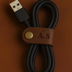 May include: Black USB cable with a brown leather strap. The strap is secured with a gold-colored rivet and embossed with the initials "A.S.". The cable is neatly coiled, suggesting organization.