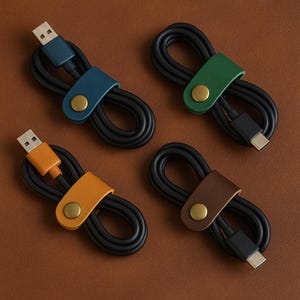 May include: Four black USB charging cables are neatly coiled and secured with leather cable organizers. The organizers are in blue, green, orange, and brown, each with a gold snap closure. The background is a warm brown.