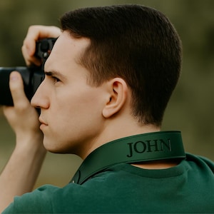 Puede incluir: Una correa de cámara verde con el nombre "JOHN" grabado en letras negras. La correa está alrededor del cuello de una persona que sostiene una cámara negra. La persona lleva una camisa verde.
