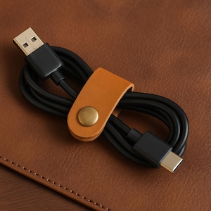 May include: A black USB charging cable with a Type-C connector, secured by a tan leather cable organizer with a brass snap. The cable is coiled on a brown leather surface.
