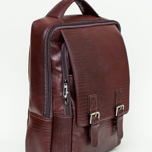 Puede incluir: Una mochila de cuero marrón con un acabado texturizado. La bolsa tiene un asa superior, una solapa frontal con dos cierres de hebilla y un bolsillo con cremallera. La mochila tiene forma rectangular.