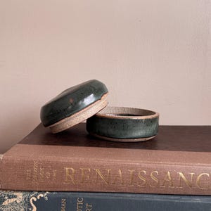 May include: Two small, dark green ceramic bowls with a speckled glaze and a tan rim are stacked on top of a stack of books. The books are brown and dark blue with gold lettering. The bowls have a rustic, handmade appearance.