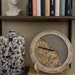 Puede incluir: Un reloj de marea de madera con un mapa de una costa. La esfera del reloj está marcada con marea alta, marea baja y marea creciente y decreciente. El reloj está sobre un soporte de madera y está sobre un libro titulado "Italia" de Ettore Pettinaroli.