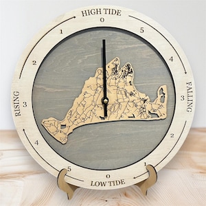 Puede incluir: Reloj de mareas de madera con marco de madera clara y centro gris. La esfera del reloj presenta un mapa de una zona costera. El texto en la esfera del reloj incluye "HIGH TIDE", "RISING", "FALLING" y "LOW TIDE". El reloj está soportado por un soporte de madera.