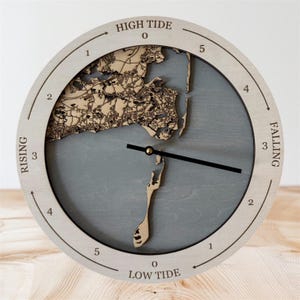 May include: A circular tide clock with a light wood frame and a grey background. The clock face features a detailed map of a coastal area. Text on the clock face includes "HIGH TIDE," "LOW TIDE," "RISING," and "FALLING."